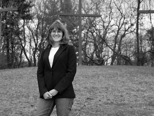 Woman in a black blazer standing outdoors with trees and crosses in the background.