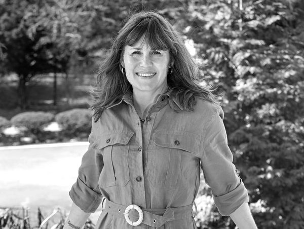Smiling woman in a belted jacket outdoors on a sunny day.
