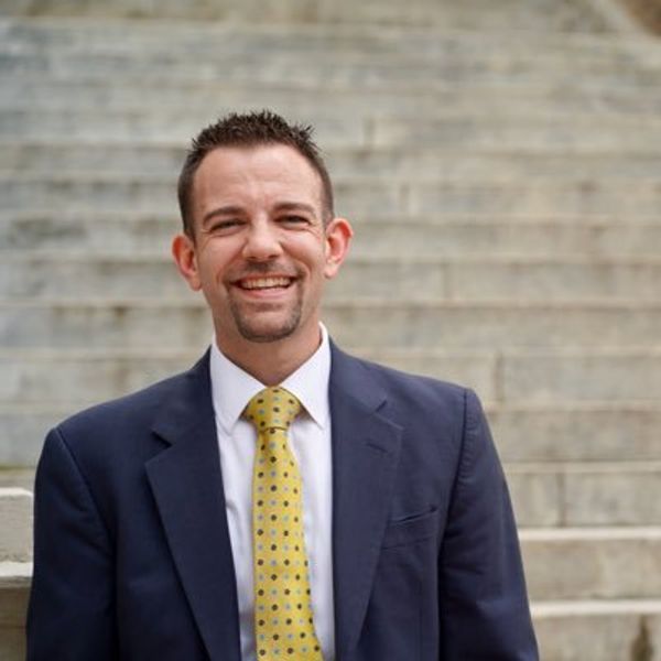 Smiling man in a suit and yellow tie standing in front of stone steps.