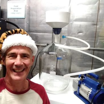 Man wearing a festive hat smiles next to a large glass container and pump in a lab.