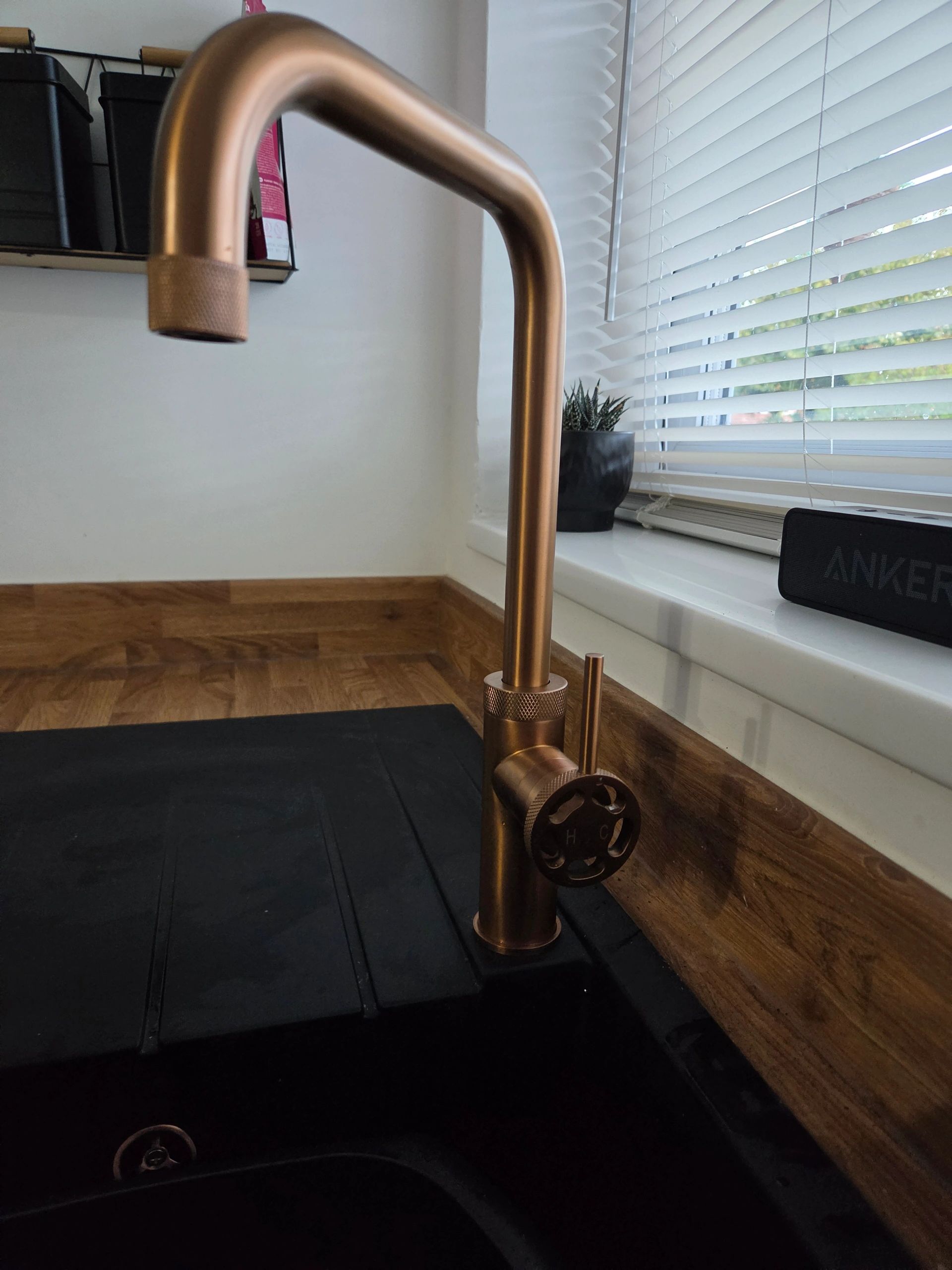 Copper-colored kitchen faucet with a black sink and window blinds.