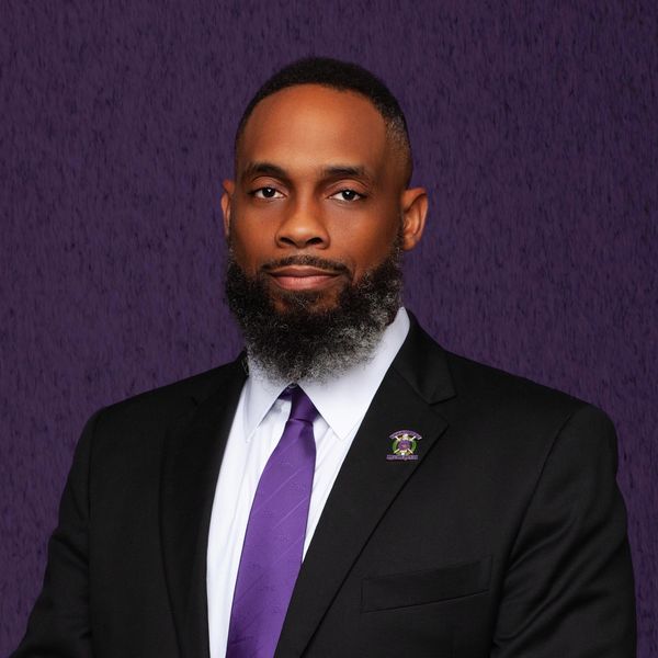 Confident man in a black suit and purple tie against a purple backdrop.