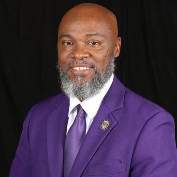 Man in a purple suit with a gray beard smiles against a black background.