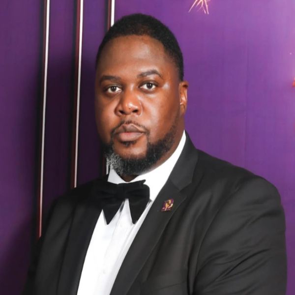 Man in a black tuxedo with a bow tie against a purple backdrop.
