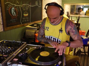 A tattooed DJ in a yellow jersey mixes vinyl records in a cozy room.