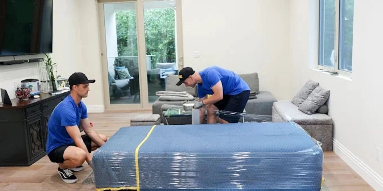 Two men in blue shirts wrapping furniture in protective plastic inside a living room.