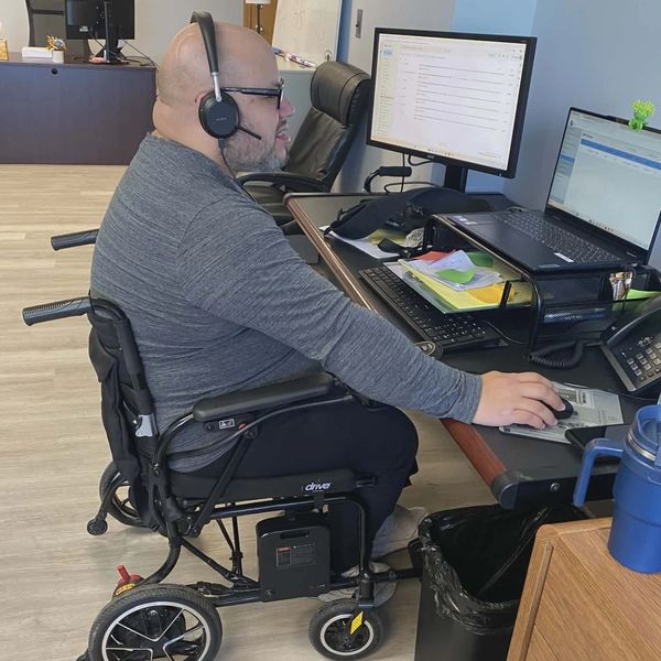 Man in wheelchair wearing headset working at dual monitors in an office.