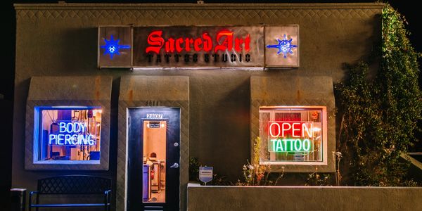 Night view of Sacred Art Tattoo Studio with neon signs for body piercing and tattoos.