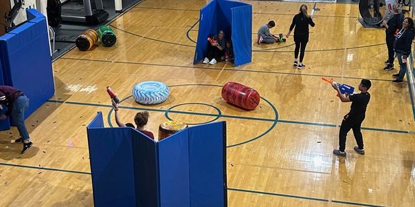 People play a Nerf gun battle game in a gym with inflatable barriers.