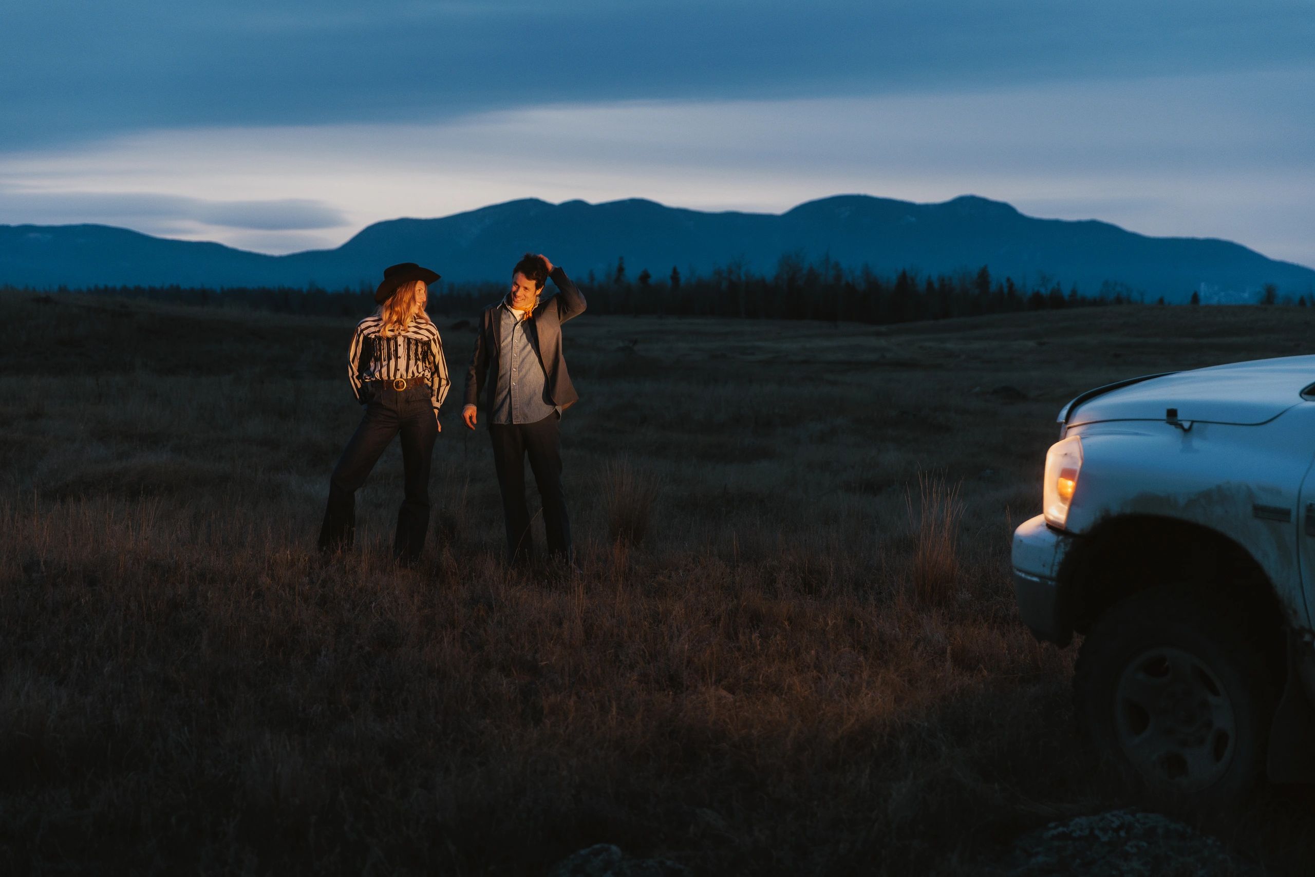 Two people standing outdoors by a truck at dusk with mountains in the background.