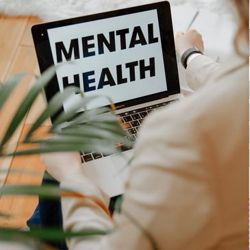 Person in brown jacket, next to a plant on laptop logging into a virtual therapy session