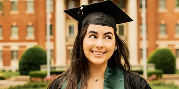 Young woman in graduation cap and gown smiling outdoors.
