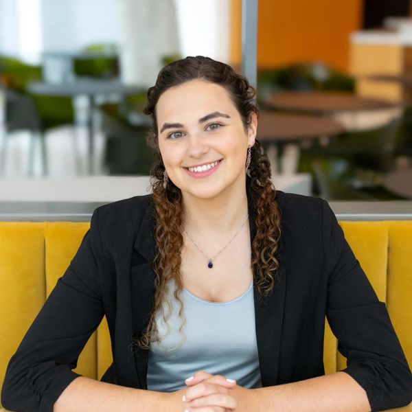 Smiling young woman with curly hair in a black blazer sitting on a yellow couch.