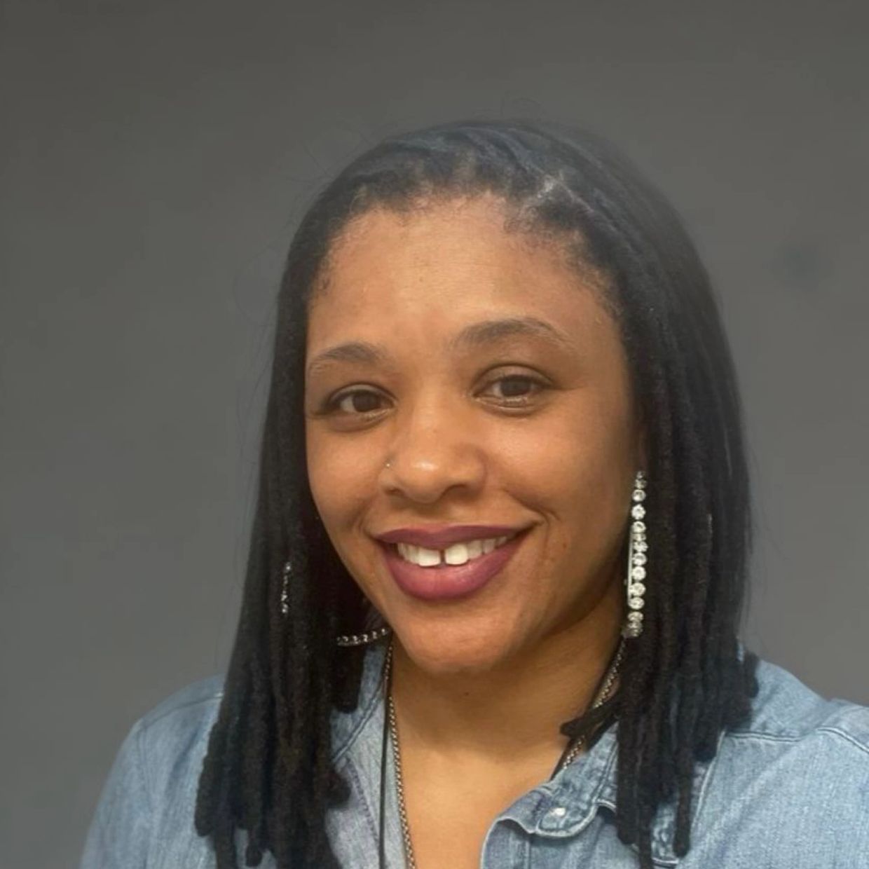 Smiling woman with dreadlocks and sparkling earrings in a denim jacket.