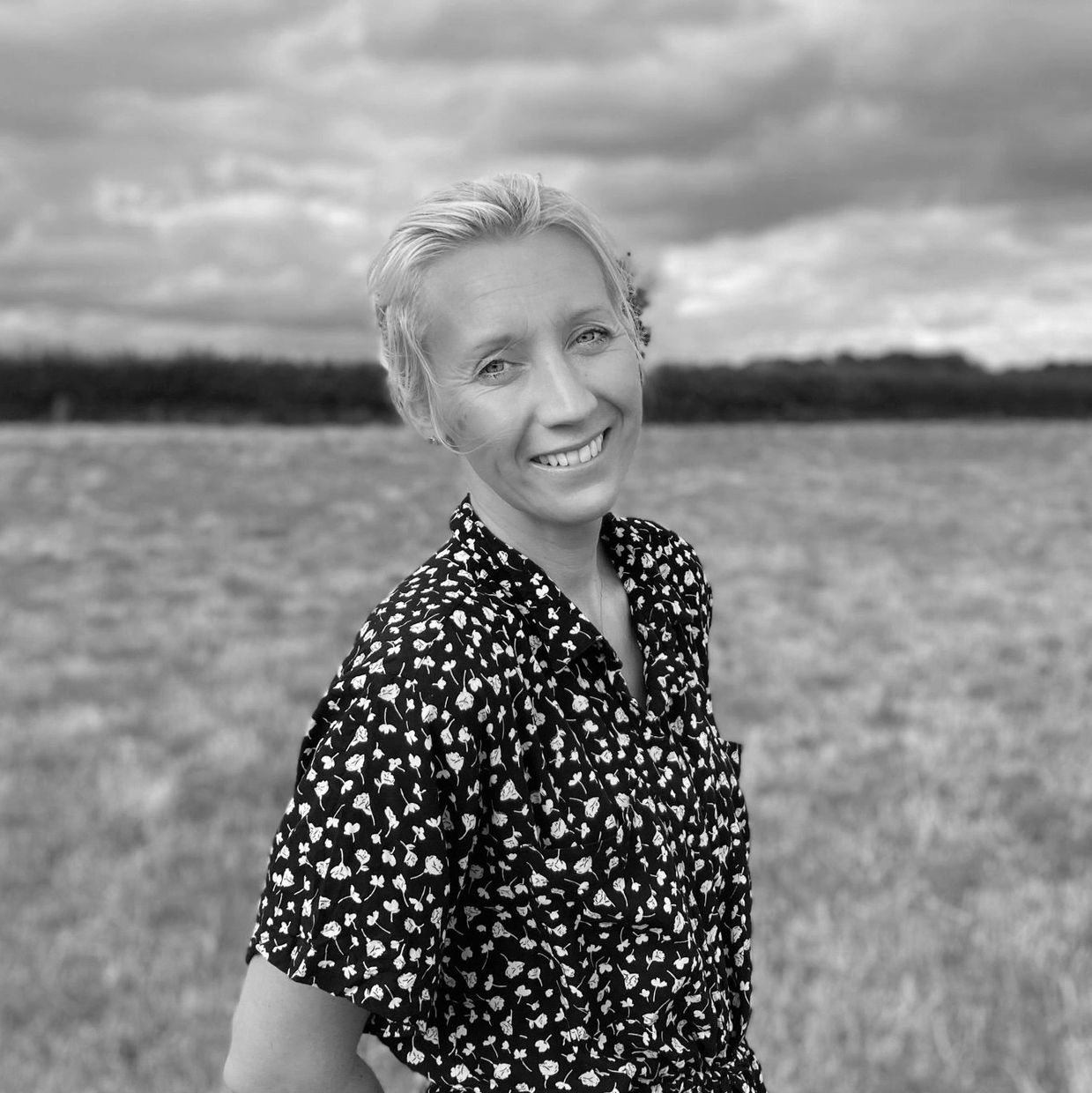 Smiling woman in a floral dress stands in a field under a cloudy sky.