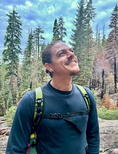 A man with a backpack smiles looking up in a forest.