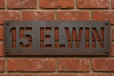 Rustic metal house number plaque reading '15 ELWIN' mounted on a red brick wall.