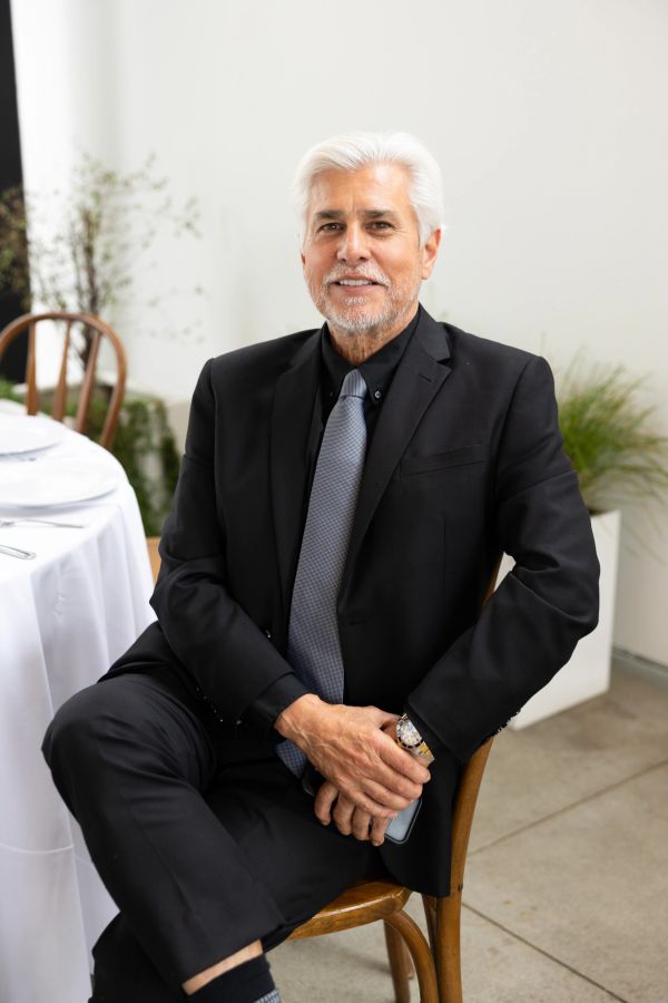 Older man in a black suit sitting confidently on a chair.