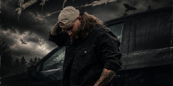 Man in dark clothing and cap stands pensively by a black vehicle under a stormy sky.