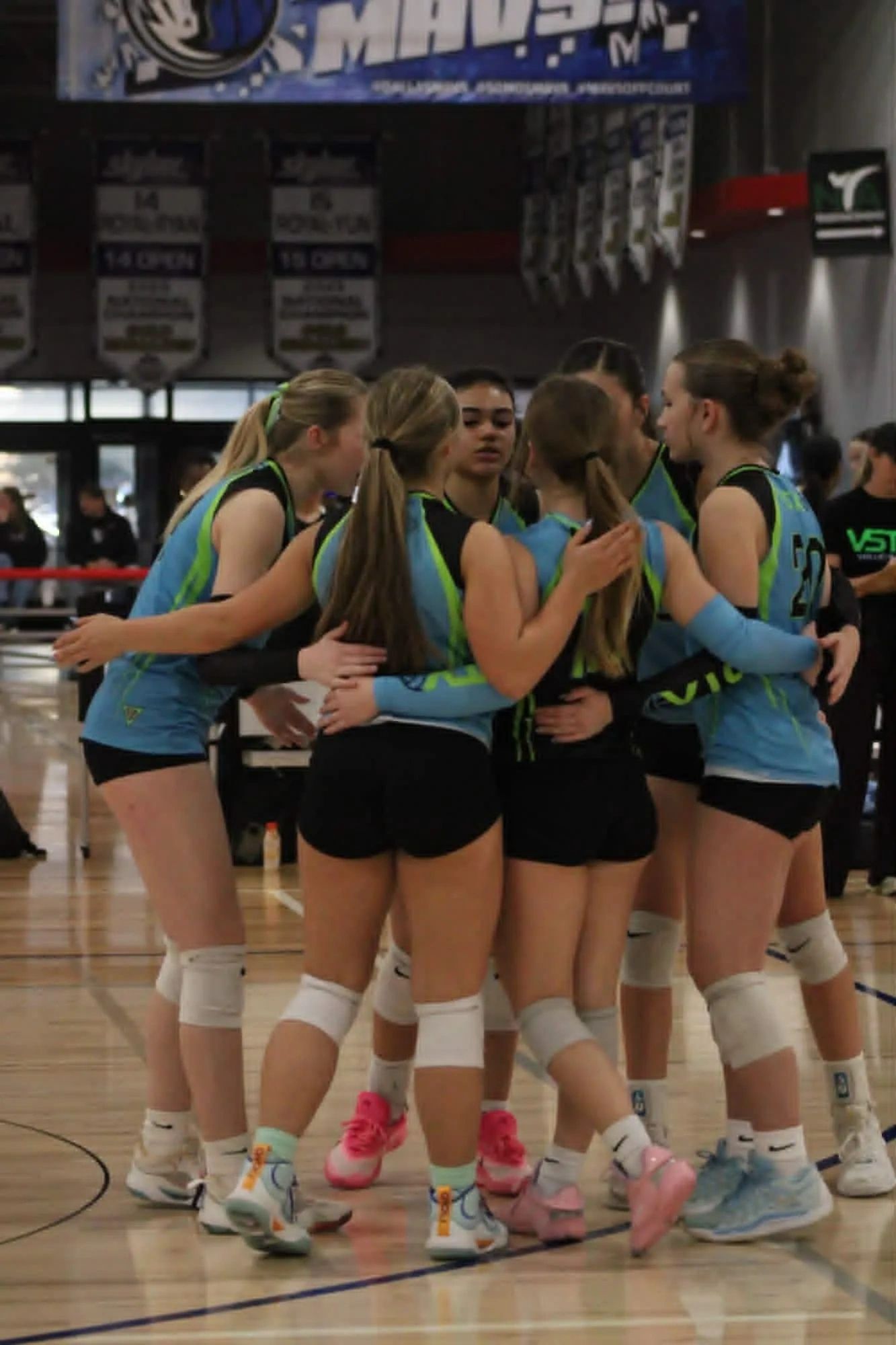 A girls' volleyball team huddling during a game in a gymnasium.