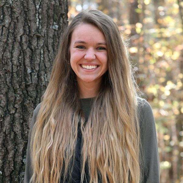 Smiling woman with long blonde hair standing by a tree in a forest.