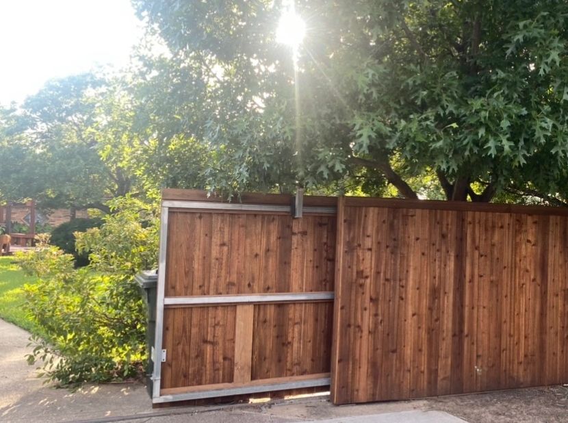 Wooden gate partially open under bright sunlight and leafy trees.