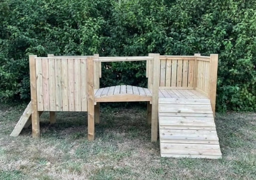 Wooden outdoor play structure with ramps and platforms in a grassy area.