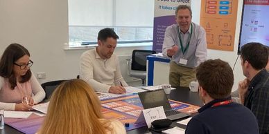 A group in a meeting room engaged in discussion around a table with posters.