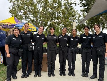 A group of nine women in matching black uniforms standing outdoors, smiling.