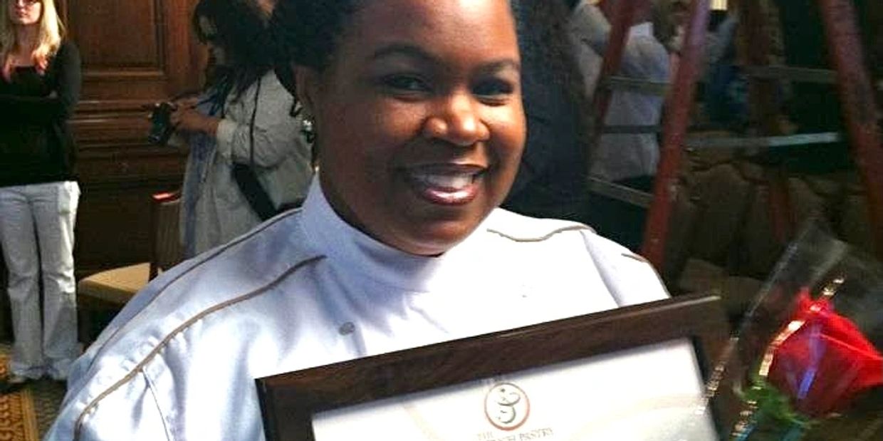 Woman in chef uniform holding a framed certificate and a red rose, smiling.