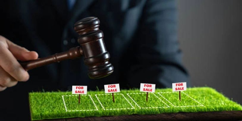 Gavel striking near miniature plots with 'For Sale' signs on grass.