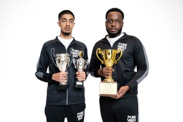 Two men holding trophies wearing matching black Pure Vibes tracksuits.
