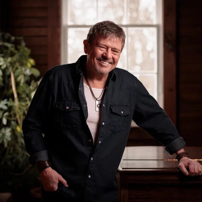 A man in a black shirt smiles warmly indoors near a window.