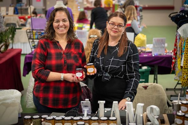 Two women at a market stall holding jars of homemade preserves.