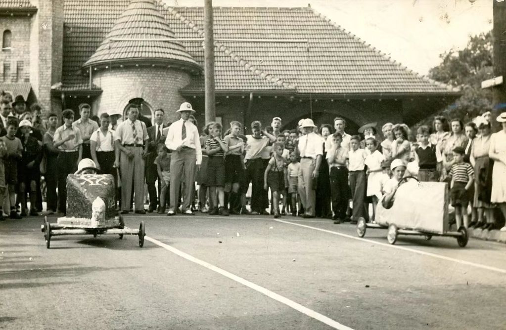 Vintage soapbox derby race with children and spectators in front of a building.