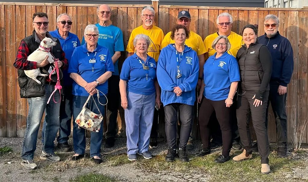 Group of people in blue and yellow shirts posing outdoors by a wooden fence, one holding a small white dog.