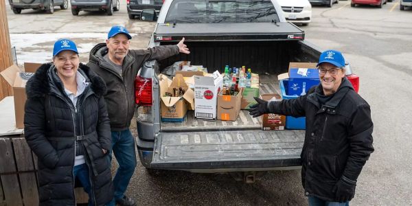 Three people with blue hats stand by a truck bed filled with boxes and bottles.