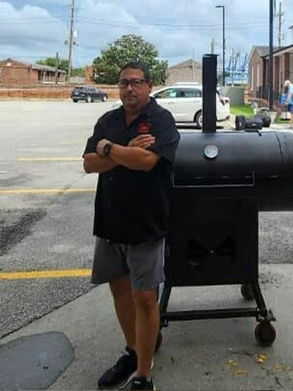 Man standing confidently next to a large black smoker grill outdoors.