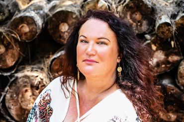 Woman with long dark hair standing in front of stacked logs.