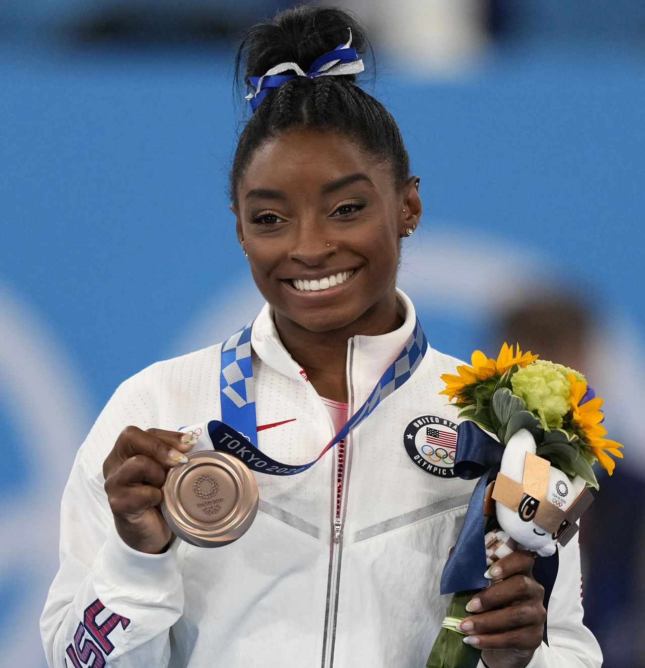 African-American woman wearing white jacket shows a medallion around her neck while holding flowers..