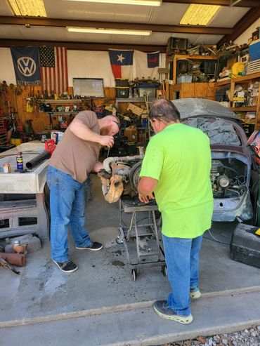 Two men working on a car engine in a cluttered garage with flags on the wall.