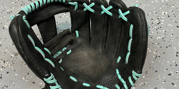 Black leather baseball glove with teal lacing on a speckled floor.
