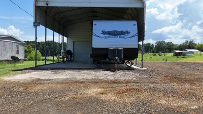 Camping trailer parked under a metal canopy in a rural area.