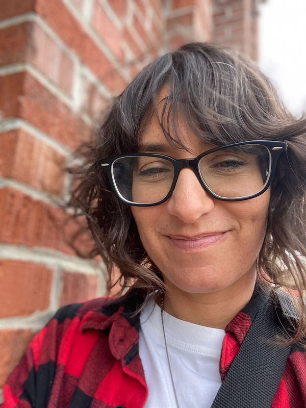 Smiling woman with glasses and curly hair in a red and black plaid shirt.