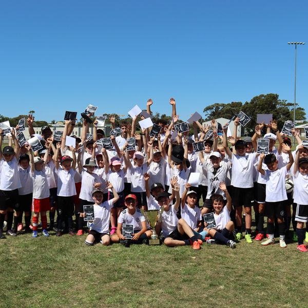 A large group of children in sportswear celebrating with certificates and a trophy outdoors.