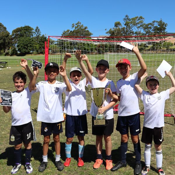 Young boys celebrating with a trophy in front of a soccer goal on a sunny day.