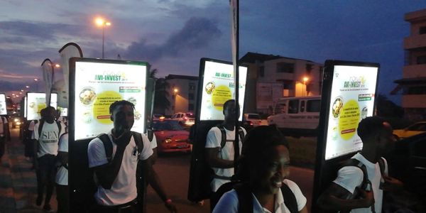 Group of people walking at dusk with illuminated advertisement boards on their backs.