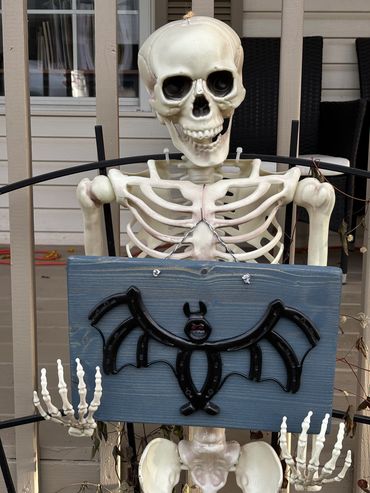 Skeleton holding a Halloween sign with a black bat decoration.