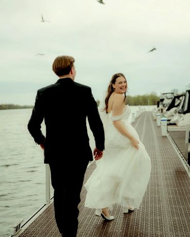 A bride and groom walking on a dock by the water, the bride smiling back.