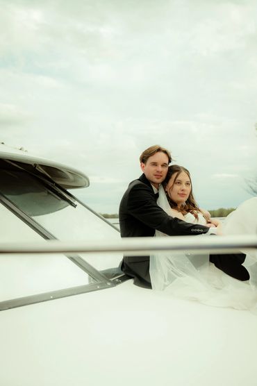 A couple in wedding attire embraces on a boat under a cloudy sky.